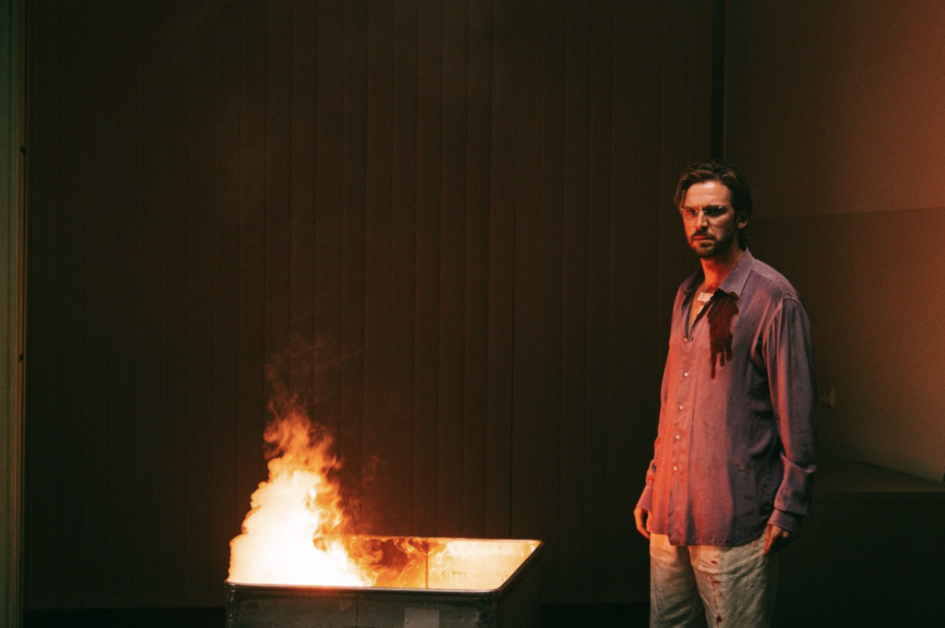 A man with a disheveled appearance and bloodstained shirt stands beside a burning dumpster in a dimly lit room.